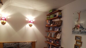 Un dormitorio con una cama, dos almohadas, una mesita de noche con libros, una estantería en la pared con libros y un póster.