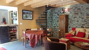 Una sala de estar acogedora con pared de piedra, escalera de madera, comedor con mesa y sillas, y sofá con cojines estampados.