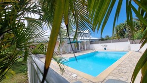A swimming pool with a stone deck, surrounded by palm trees and a white building with a blue roof. - COMFORTABLE VILLA WITH PRIVATE POOL  (SAINT FRANCOIS)