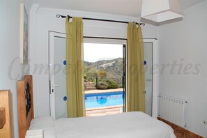 A bedroom with a bed, a door leading to a balcony with a pool, and a view of hills.