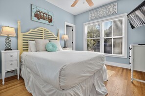 Un dormitorio con cama, mesitas de noche, ventilador de techo, ventana con persianas y una obra de arte colgada en la pared.