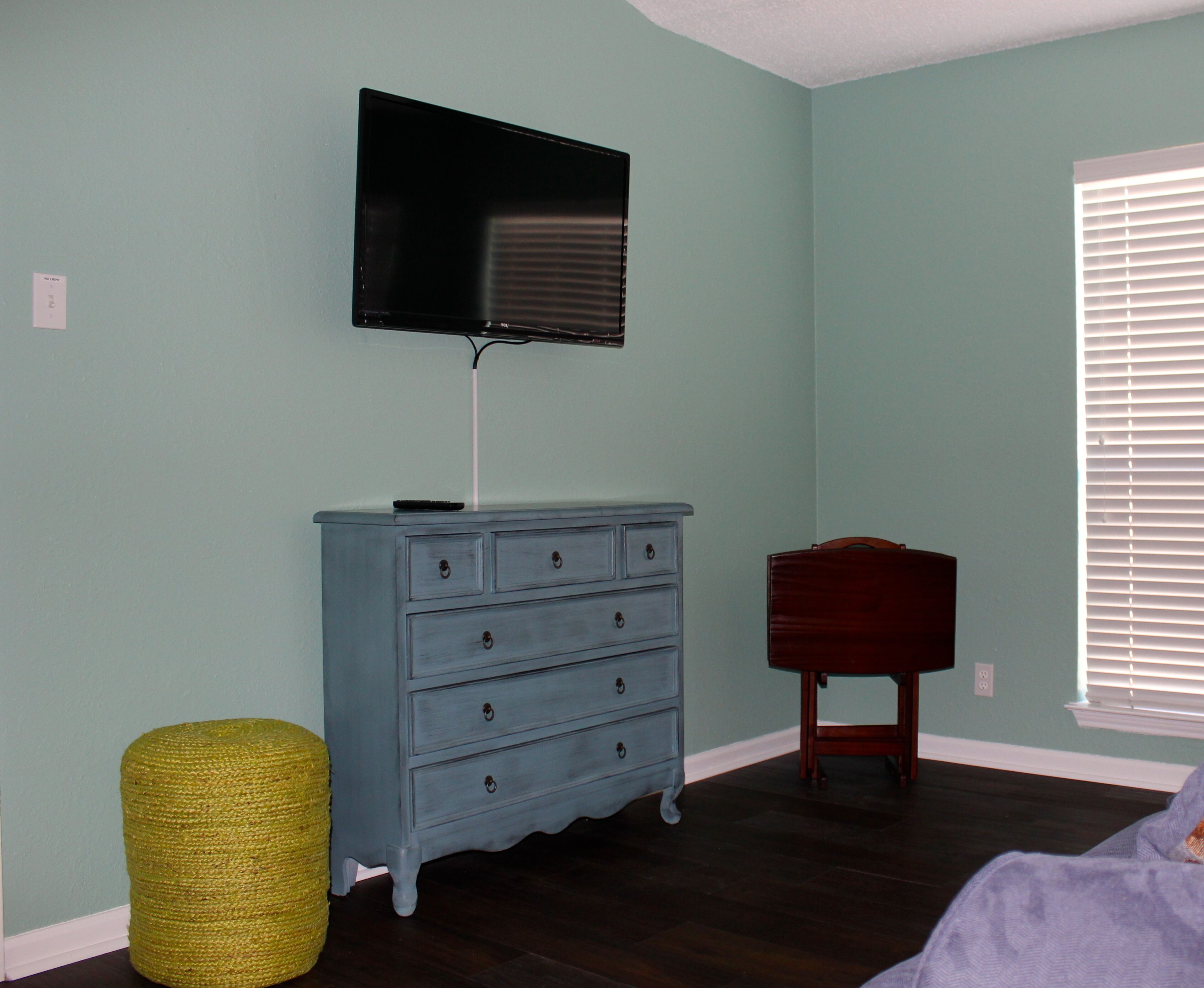 Una habitación con paredes verde azulado, un aparador de madera con cajones, un televisor montado encima, una pequeña silla de madera y una ventana con persianas.