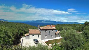 Una casa moderna de dos pisos con techo de tejas, rodeada de exuberante vegetación y vistas al mar.