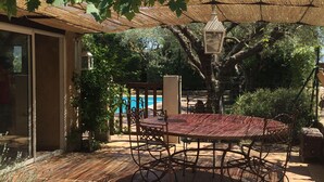 An outdoor patio with a table and chairs, a pool in the background, and a pergola providing shade.