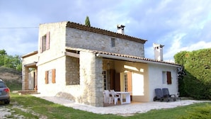 Une maison en pierre à deux étages, dotée d’un patio, d’un espace salon extérieur et d’une voiture garée à proximité.