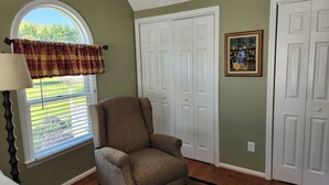 Ein Zimmer mit einem großen Fenster, einem Karomuster-Vorhang, einem Sessel und einer Tür mit einem gerahmten Bild.
