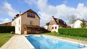 Una piscina con terraza de piedra, rodeada por un césped cuidado y una casa grande con balcón.