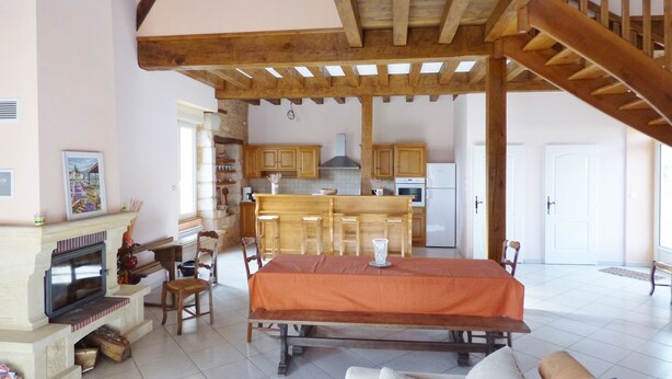 Amplia sala de estar con chimenea, una mesa de comedor y un sofá. Se ve una cocina al fondo con armarios de madera y un refrigerador.