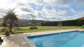 A swimming pool with a tiled deck, surrounded by a grassy area with trees and a hedge.