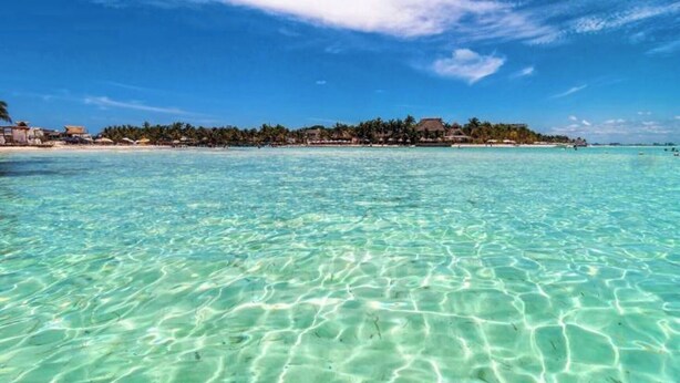 A beach with clear turquoise water and a distant view of a resort with buildings and palm trees. - Villa #3 - Isla Retreats | Tropical Gardens + Pool (Isla Mujeres)