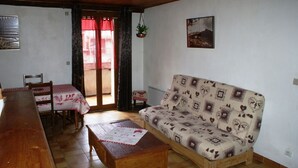 Ein gemütliches Wohnzimmer mit einem Holzsofa, einem Esstisch mit Stühlen und einem großen Fenster mit roten Vorhängen.