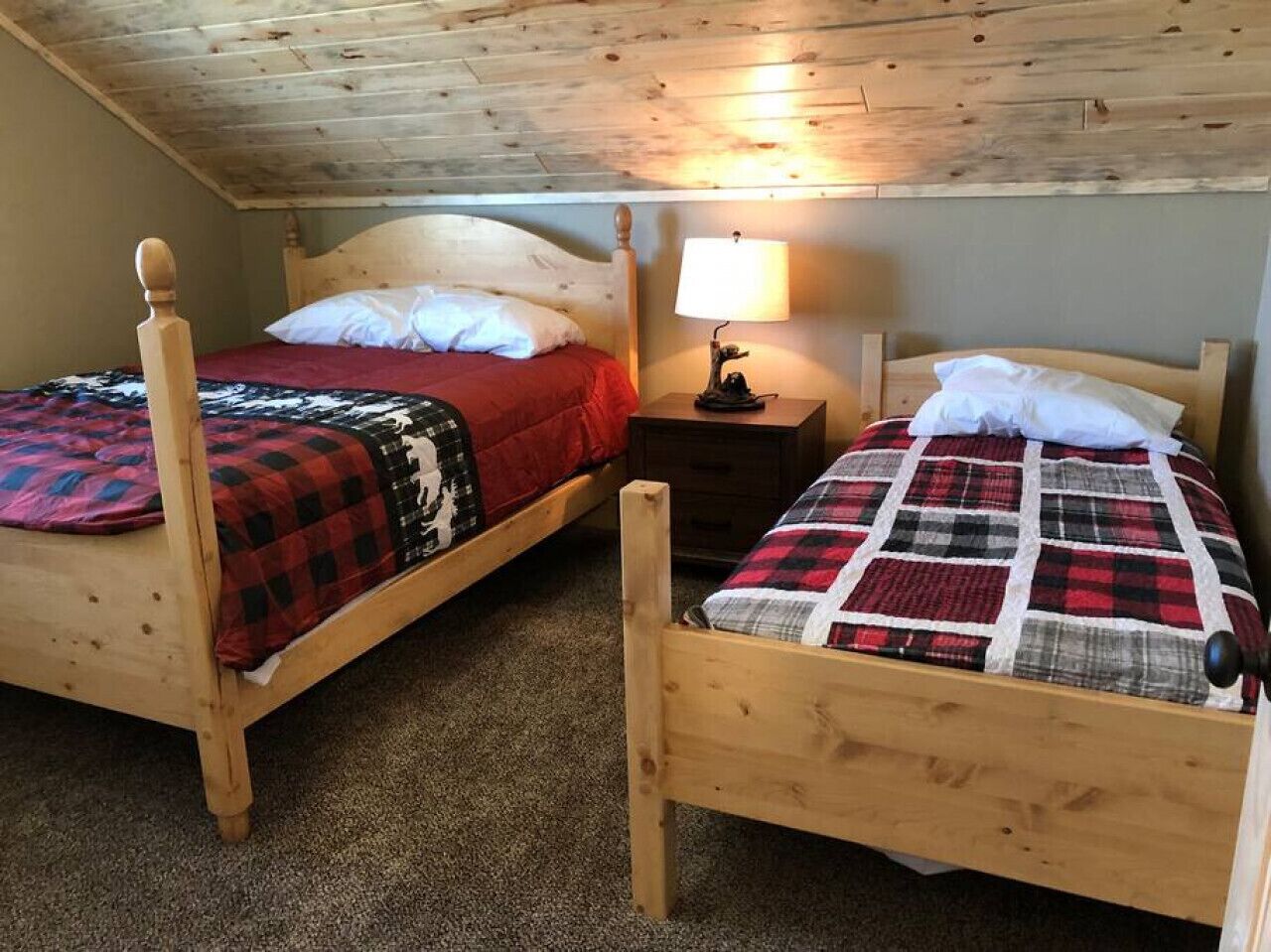 To senge af træ med rødt og sort ternet sengetøj, et natbord med en lampe, og et træloft.