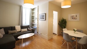 Ein Wohnzimmer mit Sofa, Couchtisch und Esstisch mit Stühlen. Es gibt ein Fenster mit Vorhängen, ein Bücherregal und einen Blumentopf.