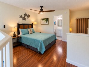 Une chambre à coucher avec un lit, une table de chevet, un ventilateur de plafond, une salle de bain visible à travers une porte ouverte et un plancher en bois.