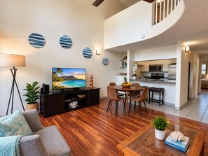 A modern living room with a flat-screen TV, wooden flooring, a dining area with a table and chairs, and a kitchen in the background.