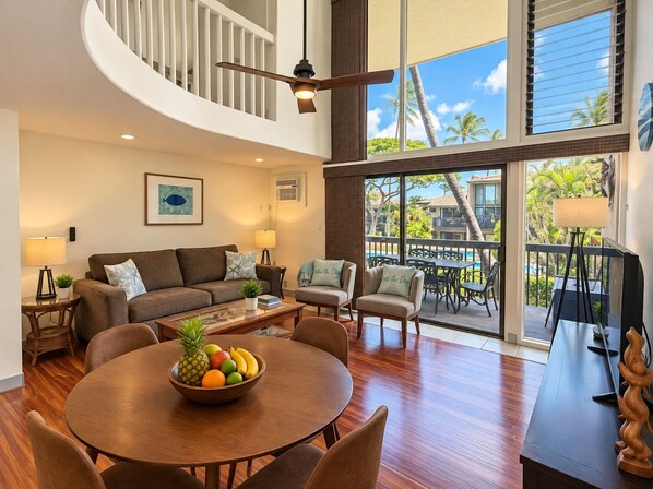 A spacious living room with a wooden dining table, comfortable seating, a ceiling fan, and large windows offering a view of the outdoors.