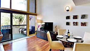A spacious living area with a large TV, dining table, and chairs. There is a balcony with a view of greenery. - Where Families Gather Kihei 2BR Loft Condo Heated Pool Hot Tub Near Beach (Kihei)