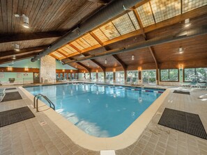 Une piscine intérieure dotée d’un plafond en bois et de murs en pierre, entourée de chaises longues et offrant une vue dégagée sur l’extérieur.
