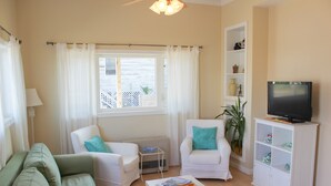 A living room with a green sofa, two white armchairs with teal cushions, a white coffee table with magazines, a white entertainment unit with a TV, and a window with white curtains. - Beach Odyssey (Saco)