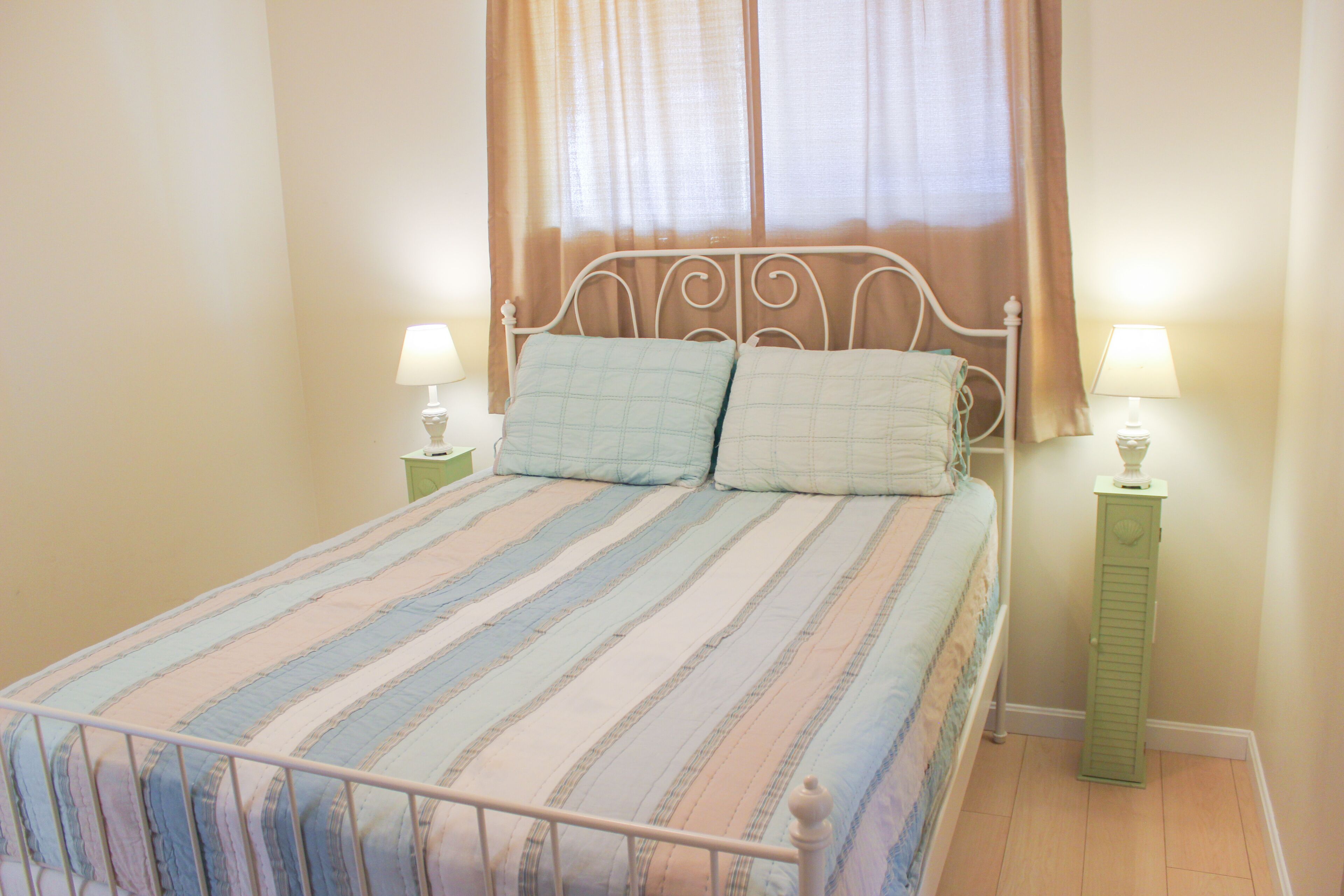 Uma cama bem arrumada com roupa de cama listrada, duas mesas de cabeceira com abajures e uma janela com cortinas.