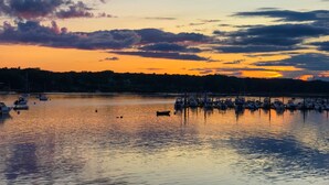 A marina with boats docked at the pier, calm water reflecting the sunset, and a forested shoreline in the background. - Beach Odyssey (Saco)