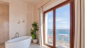 A modern bathroom with a freestanding bathtub, a potted plant, and a large sliding glass door leading to a balcony with a view of the sea.