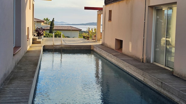 Ein Schwimmbad mit Holzterrasse, einer darüberliegenden Pergola und Blick auf ein entferntes Gebäude und die Landschaft.