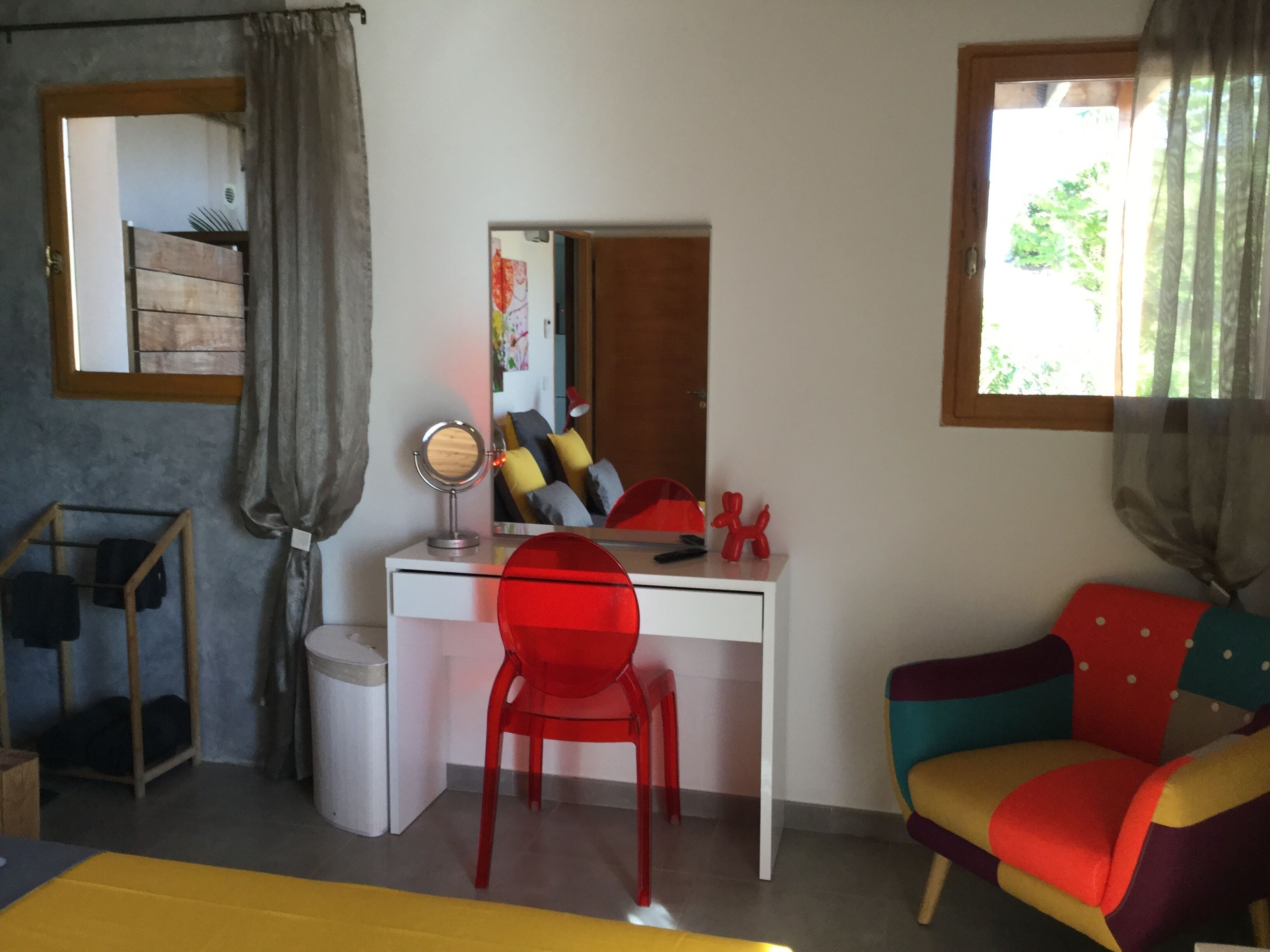 A room with a white desk, a red chair, a colorful sofa, a small round mirror, and a window with curtains.