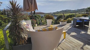 Eine Holzterrasse mit Gartenmöbeln, darunter ein weißer runder Tisch und Stühle, sowie Kübelpflanzen.