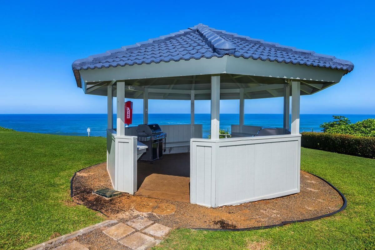 A gazebo with a tiled roof, a grill, and a seating area with a table and chairs, overlooking the ocean.