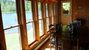 A dining area with a large table and chairs, wooden walls, and large windows overlooking a body of water. - Adirondack Beauty! 110 ft Lake Frontage On Brant. Lake - Beach; Kayaks, Canoes (Greig)