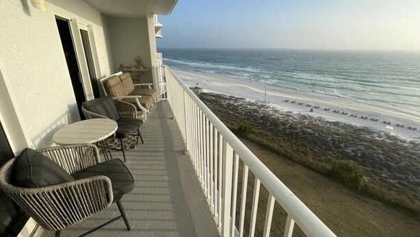 A balcony with wicker chairs and a table, overlooking a beach with a rocky shoreline and calm sea. - Beachfront cozy 2 bedroom 2 bath (Panama City Beach)