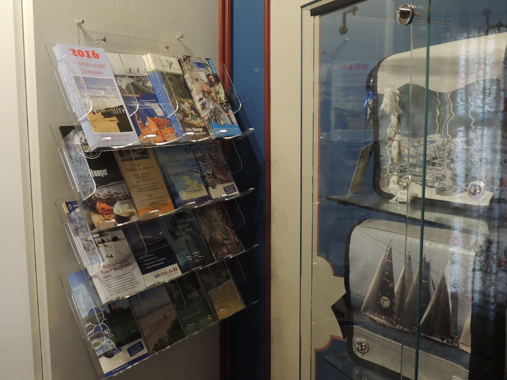 Una vetrina espositiva con un modellino di nave d'epoca e una rastrelliera a parete per brochure con diverse informazioni su viaggi ed eventi.