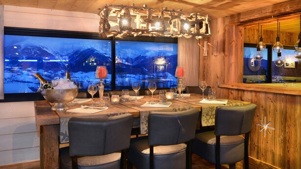 Un restaurant au décor intérieur en bois, avec une grande fenêtre offrant une vue sur les montagnes enneigées, et un coin bar agrémenté de luminaires suspendus.