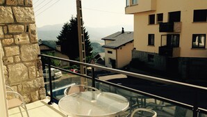 Un balcon avec une table et des chaises en verre, donnant sur une rue bordée d’immeubles et offrant une vue lointaine sur les montagnes.