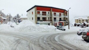Un immeuble à plusieurs étages, avec un toit rouge et une façade en pierre, entouré d’un sol enneigé et de voitures garées.