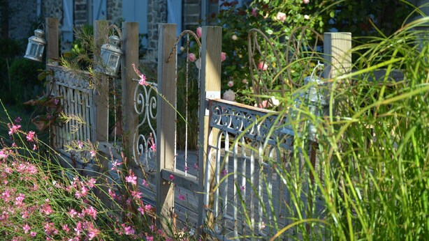 Una rústica puerta de madera con detalles de metal decorativo y faroles, rodeada de exuberante follaje y flores rosadas.