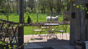 Un área de comedor al aire libre con encanto, con sillas de metal y una mesa, rodeada por una valla de madera y un campo cubierto de césped con una oveja al fondo.