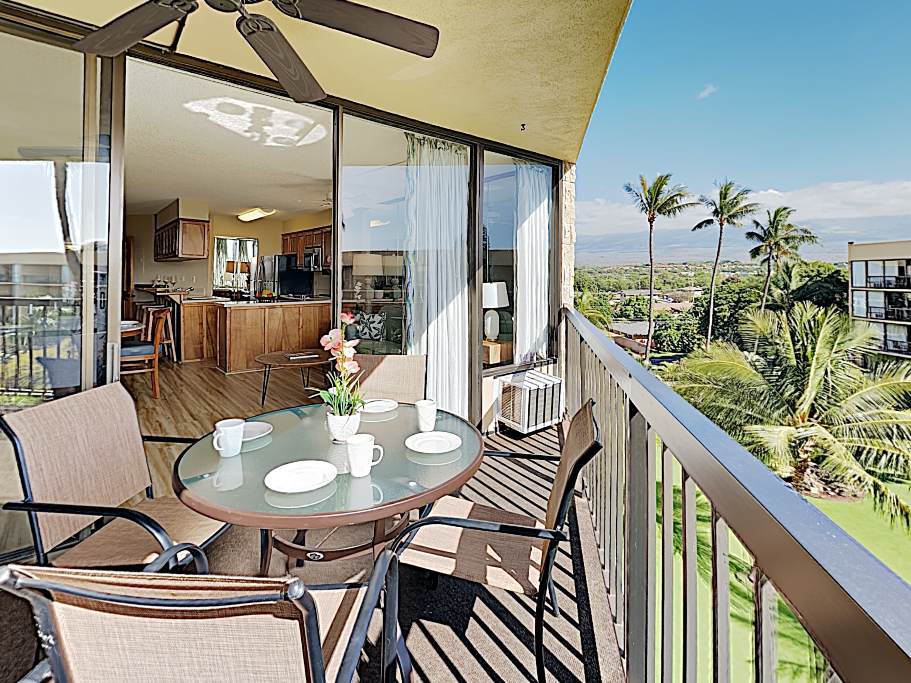 A balcony with a table and chairs, a ceiling fan, and a view of a lush landscape with palm trees.