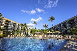 A hotel pool area with multiple lounge chairs and umbrellas, surrounded by palm trees and a clear blue sky.