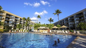 Zwembadgedeelte van een hotel met meerdere ligbedden en parasols, omgeven door palmbomen en een helderblauwe lucht.