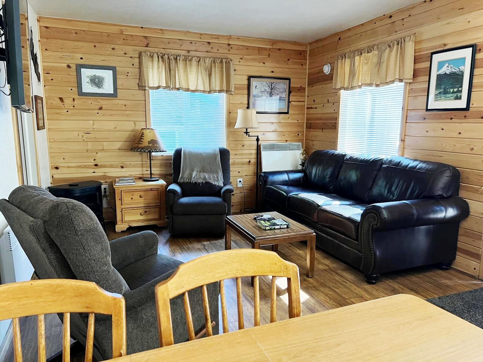 Ein gemütliches Blockhauskamin mit Ledersofa, Holzesstisch, einem Stuhl, einem kleinen Beistelltisch und einer Lampe.