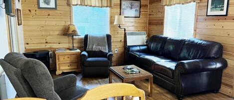 Ein gemütliches Blockhauskamin mit Ledersofa, Holzesstisch, einem Stuhl, einem kleinen Beistelltisch und einer Lampe.