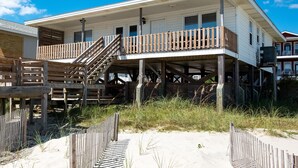 Una casa sulla spiaggia con una veranda in legno, una scala che conduce a un balcone e una spiaggia sabbiosa in primo piano.