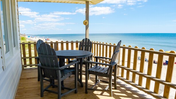 Un terrazza in legno con tavolo e sedie, affacciata su una spiaggia con una recinzione in legno e un cielo sereno.