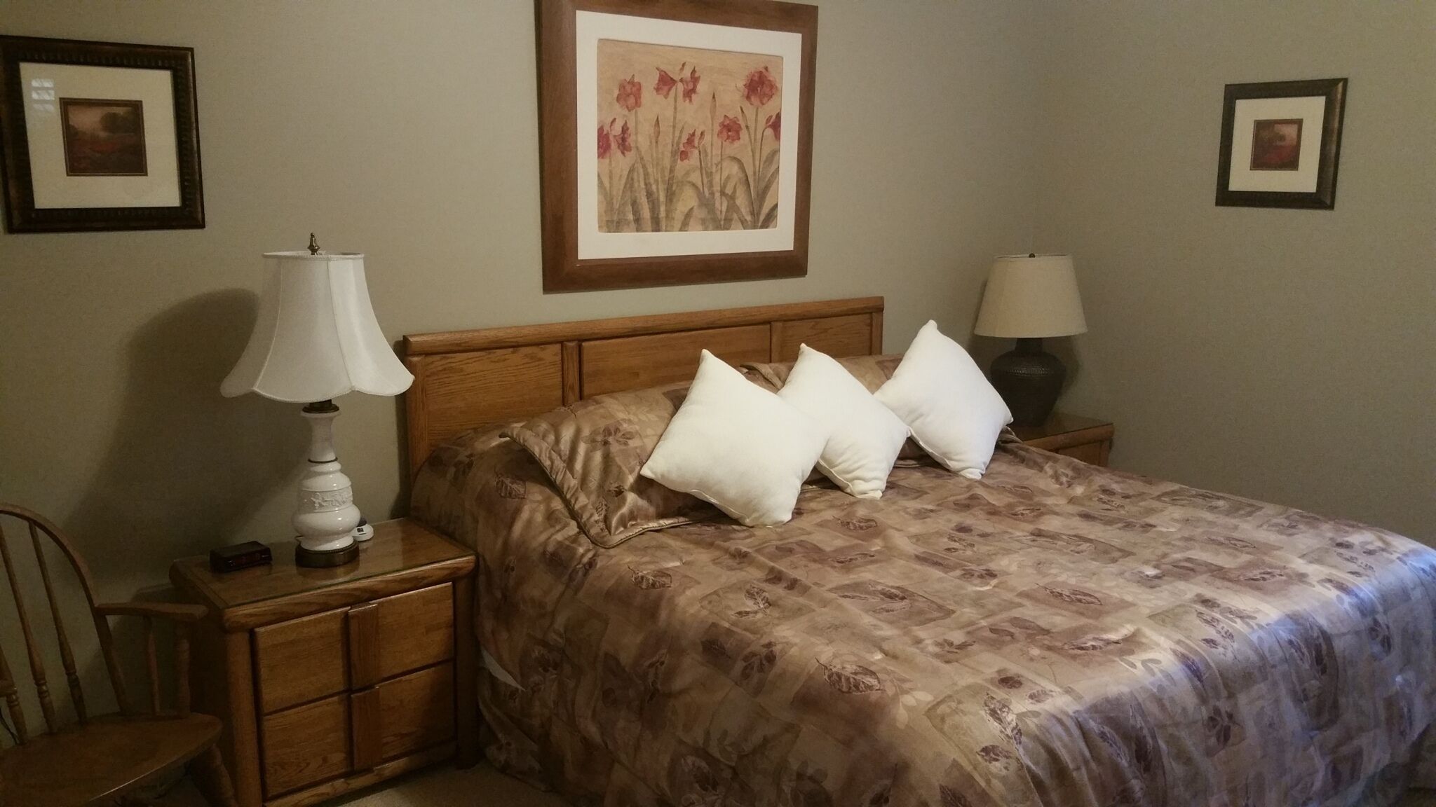 Una cama bien hecha con una colcha estampada de flores y dos almohadas blancas, una mesita de noche de madera con una lámpara, y un cuadro en la pared.