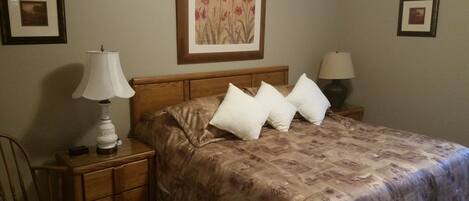Una cama bien hecha con una colcha estampada de flores y dos almohadas blancas, una mesita de noche de madera con una lámpara, y un cuadro en la pared.