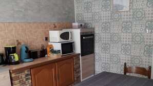 Une cuisine compacte équipée d’un micro-ondes, d’un grille-pain et d’une machine à café sur le plan de travail. Un lit recouvert d’une couverture grise se trouve au premier plan.