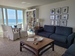 A living room with a view of the ocean, a bookshelf, a coffee table, and a couch.