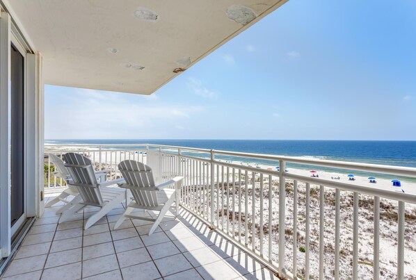 A balcony with two Adirondack chairs, a white railing, and a view of the beach and ocean.